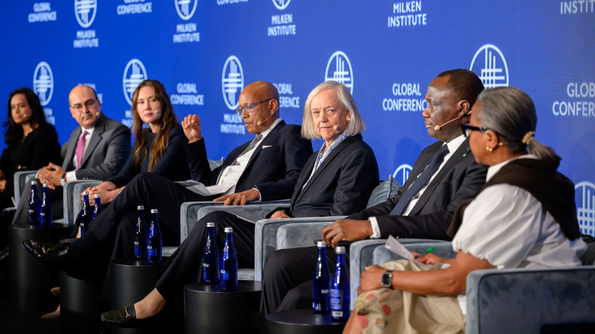 Panel discussion with six people seated on stage against blue branded backdrop